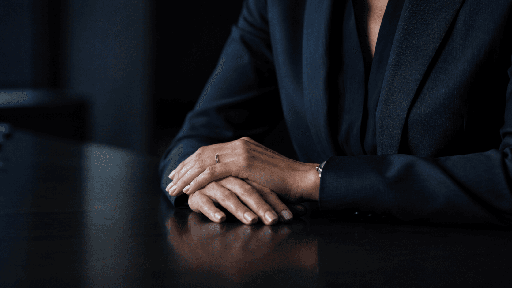 Hands in a navy suit resting on a dark, reflective table with a silver ring.